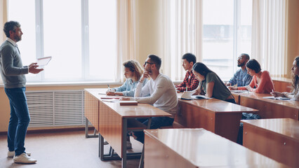 Male professor in casual clothes is talking to group of students sitting at tables in classroom and making notes. Large lecture hall with desks, chairs and large windows is visible. - obrazy, fototapety, plakaty