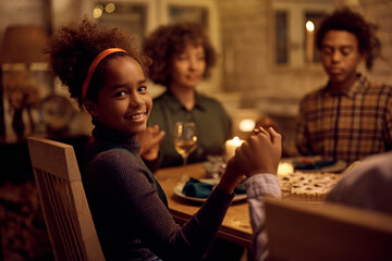 Happy black girl saying grace during family dinner on Thanksgiving and looking at camera. - Powered by Adobe