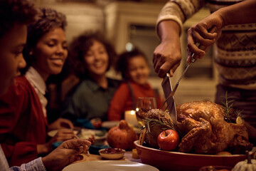 Close up of black father carving roasted turkey during Thanksgiving meal at dining table.