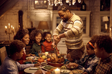 Fototapeta na wymiar Happy black man carving Thanksgiving turkey during family meal at dining table.