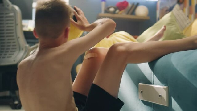 Focused young boy with a bruised arm do situps on the carpet in his room. Abs exercise. Holds a ball in his hands. Determined schoolboy for a soccer match. Preparations, fitness, sports training.