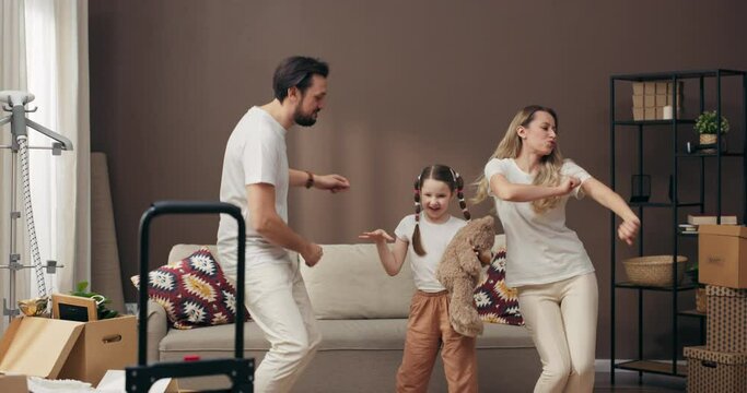 Modern family concept father with mother and daughter dancing in new spacious apartment satisfied homeowners standing in the middle af flat full of cardboxes with potted plants and household sutff .