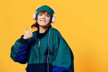 a joyful woman in a green jacket and a hat in the style of the 90s enjoys music standing in pink headphones on a yellow background showing a thumbs up