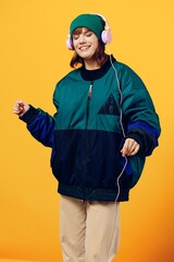 a happy, joyful woman in a green jacket and hat enjoys music while standing in pink headphones on a yellow background dancing. Vertical photography