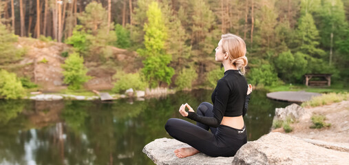 Beautiful young woman meditating near lake in forest