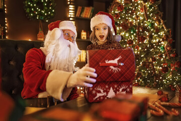 Fototapeta na wymiar Santa Claus surprising little girl while open magical gift box near Christmas tree. New Year concept