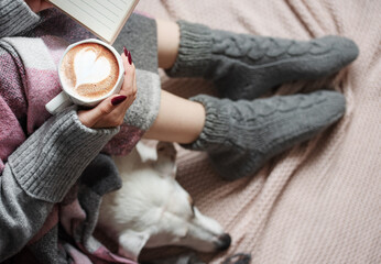 Cozy home, woman covered with warm blanket, drinks coffee,  sleeping dog next to woman.