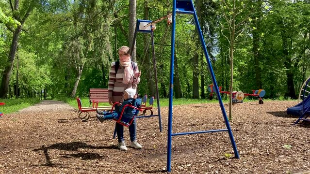 Young mommy is swinging her toddler in town city park. The baby is on the swing in sunny autumn day. Fa,ily holidays on the playground. Happy childhood.