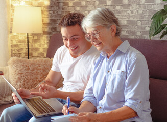 Smiling young boy sitting on sofa at home helping senior grandmother surf with laptop on the web...