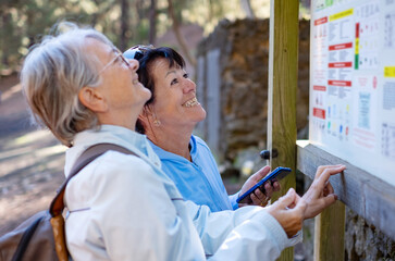 Cheerful senior couple of females friends in mountain excursion looking the right direction on the map enjoying healthy lifestyle in nature