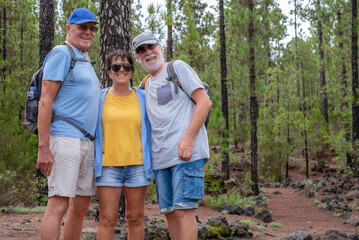 Cheerful group of senior people with caps and backpack in mountain excursion looking at camera smiling, three caucasian retired enjoying healthy lifestyle in the forest