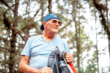 Portrait of senior caucasian man standing in the woods ready for mountain excursion. Retired male with hat sunglasses and backpack enjoying healthy lifestyle