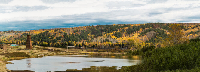Panoramic autumn landscape. Village and water tower on the shore of the lake. There is a mixed forest on the mountains, the leaves turned yellow. Beautiful sky with clouds. Nature of Eastern Siberia.