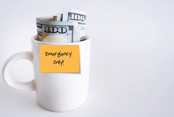 White ceramic cup with cash dollars money with orange paper stick note written EMERGENCY ONLY, isolated on white background. smart financial planning to save money for contingency fund - for rainy day