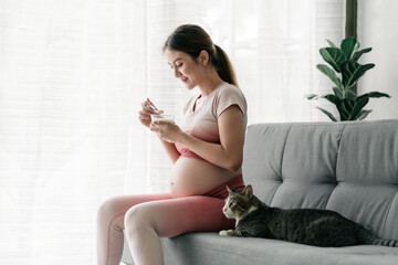 Pregnant asian woman in exercise outfits is eating yogurt on the couch with her lovely kitty...