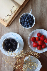 Vintage tea set filled with blueberries, blackberries and strawberries, open book, reading glasses and decorative flowers on the table. Flat lay.