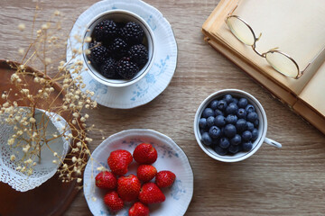 Vintage tea set filled with blueberries, blackberries and strawberries, open book, reading glasses and decorative flowers on the table. Flat lay.