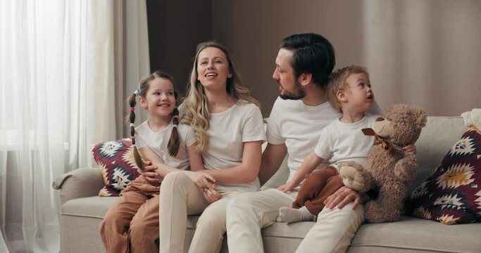 Happy family concept moving in new apartment sitting on cozy sofa relaxing discussing how to furnish decorate everething. Carton boxes full of potted plants and home stuff.