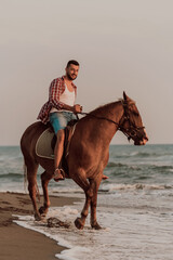 A modern man in summer clothes enjoys riding a horse on a beautiful sandy beach at sunset. Selective focus 