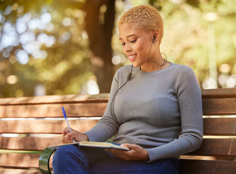 Woman, earphone and writing in book, diary or journal in a park outdoor with a happy smile on a bench. Latino girl write idea, goals and vision in a notebook in summer while relax or smiling alone