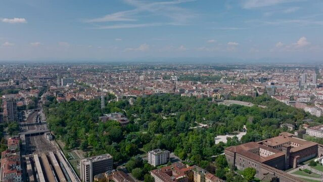Forwards fly above town. Aerial view of green deciduous trees in large public park and buildings in surrounding boroughs. Milano, Italy