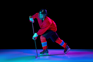 Training of male ice hockey player wearing hockey jersey, uniform and sports helmet in motion isolated over dark background in neon light