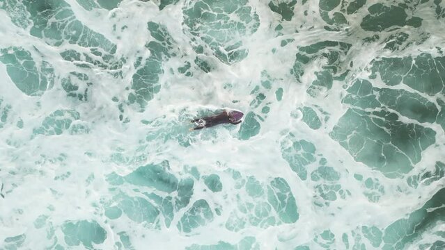 Aerial top down shot of a surfer paddling through thick white foam and over a swell of an incoming wave as he heads back out to the backline, Suances, Spain