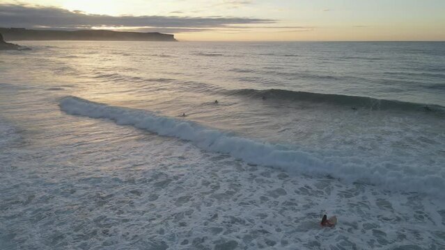 Aerial drone shot tracking backwards over a group of recreational surfers enjoying the last light as the beautiful golden sunsets in the distance over the Cantabrian Sea, Spain