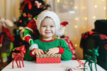 Fototapeta na wymiar Happy little smiling girl with Christmas gift box. Festive little toddler girl opening a gift at home in the living room. Christmas, holidays and childhood concept. 