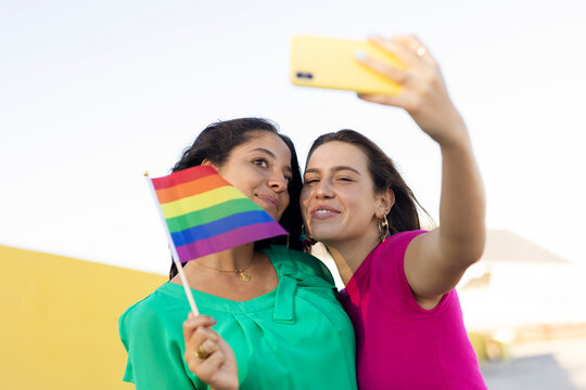  A beautiful lesbian young couple embraces and holds a rainbow flag. Girls taking selfie photo