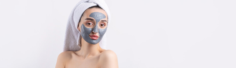 A woman with a gray clay cosmetic mask in a white towel on her head on a white background.