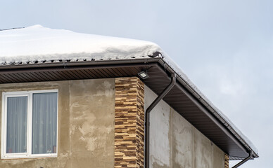 Close up view of roof slope with a lot of snow, rain gutter, downpipe and light for the motion sensor mounted on soffit of building