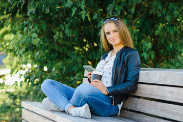 Obraz premium A smiling modern young woman sitting on bench in green park on summer day and talking on smartphone with coffee cup.Girl sitting with a phone and coffe at park.Klaipeda,Lithuania.09-02-2022.