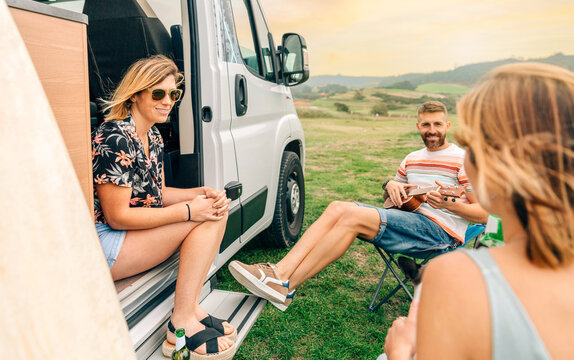 Group of friends having fun talking and playing ukulele in front of their camper van