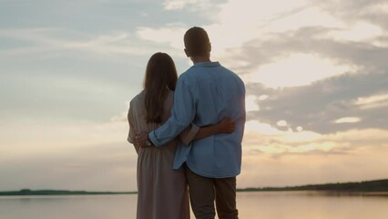 Back view of Caucasian young happy married couple standing outdoor near big lake looking at beautiful sunset and hugging. Boyfriend and girlfriend in love on walk near water. Nature. Romantic concept - Powered by Adobe