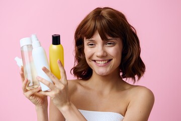 a happy young woman stands on a pink background, wrapped in a white towel and holding a set of jars with facial care cosmetics, smiling broadly and squinting her eyes