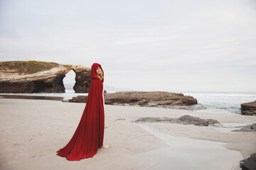 A beautiful woman in a long red cape is standing with her eyes closed on a beautiful rocky beach