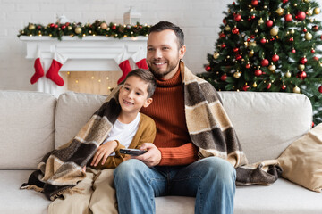 cheerful father and son sitting on couch under warm blanket and watching tv near decorated christmas tree