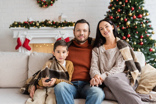 Boy With Remote Controller Near Happy Family Watching Tv On Couch Under Warm Blanket Near Blurred Christmas Tree