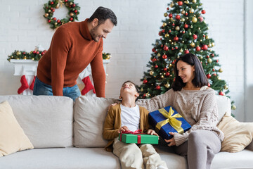 happy boy looking at dad while sitting on couch with christmas presents and smiling mom