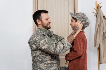side view of bearded man putting military cap on cheerful son meeting him at home