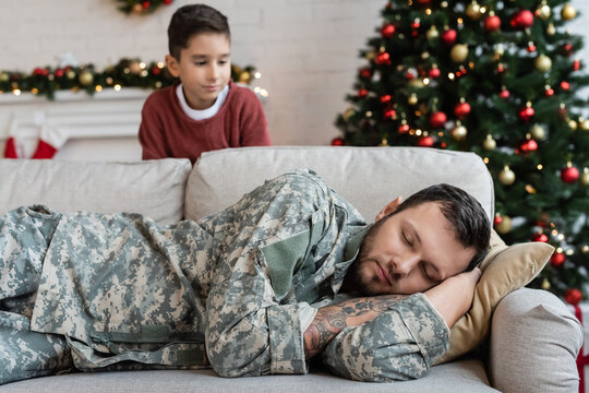 Blurred Boy Looking At Exhausted Dad In Camouflage Sleeping On Couch In Living Room With Christmas Decoration