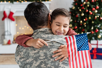 cheerful boy with closed eyes and usa flag embracing dad in camouflage at home