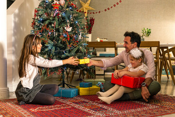 A father is sitting on the floor next to the Christmas tree with his daughters