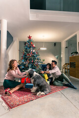 A mother and father sitting with their daughter on the floor next to the Christmas tree and exchanging Christmas presents. The living room is decorated in the spirit of the upcoming holidays.
