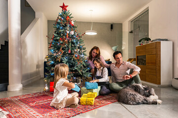 A mother and father sitting with their daughters and dog on the floor next to the Christmas tree and exchanging presents. The living room is also decorated in the spirit of the upcoming holidays.