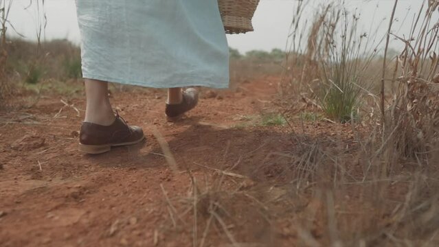 Close-up of a woman in a blue dress with a basket in her hand walking across a dirt field