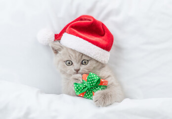 Playful kitten wearing red santa's hat hugs gift box under white warm blanket. Top down view