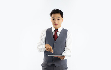 Young asian businessman in grey vest suit white shirt red tie holding tablet touching screen looking camera white background.
