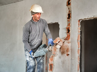 Worker in protective helmet with perforator completing deconstruction of rest of brick wall inside...